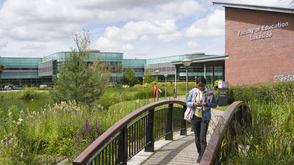 Student walks across the bridge next to the lake looking at her phone with the Faculty of Education building in the background