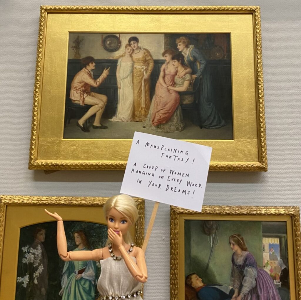 A barbie doll is positioned in front of a piece of artwork. The doll holds a placard that reads: 'A mansplaining fantasy! A group of women hanging on every word. In your dreams!'