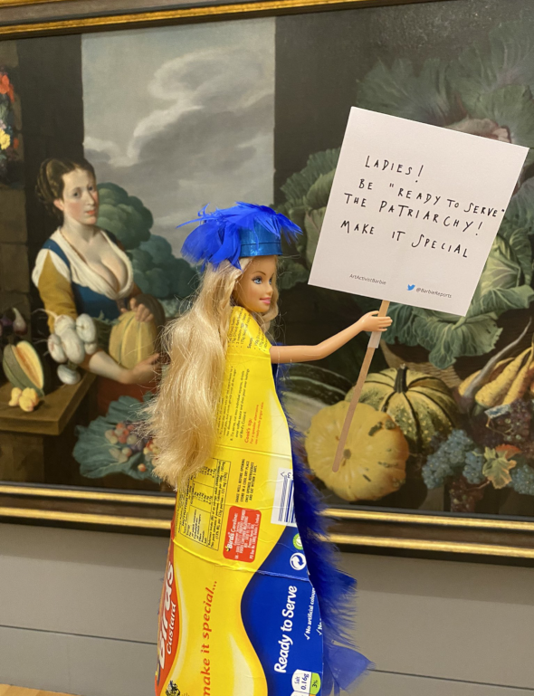 A barbie doll wearing a ready-to-serve custard wrapper stands in front of a painting. The doll holds a placard saying 'Ladies! Be ready to serve the patriarchy! Make it special'.