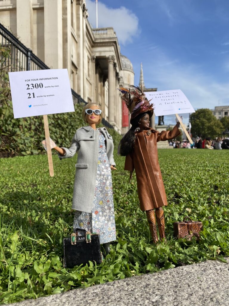 Two barbie dolls stand outside the national gallery. One holds a placard that says 'For your information. 2300 works by men. 21 works by women.' The other holds a placard that says 'Welcome to the Patriarchal Palace of Painting!'