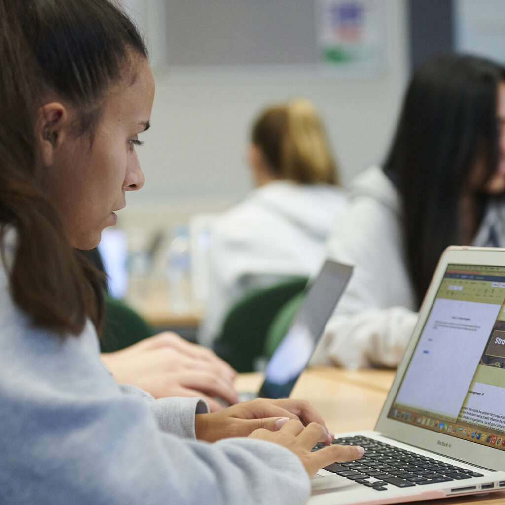 A student focuses on her laptop in a classroom setting, with other students around her also engaged in their studies.