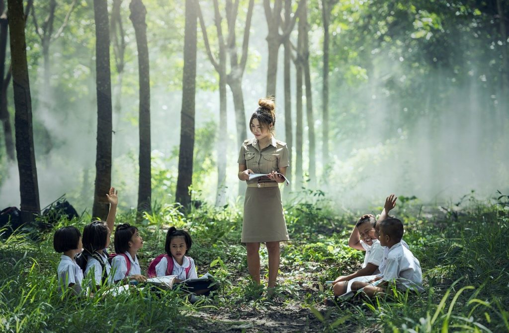 A teacher holding a notebook, standing in woodland. Her six pupils are sat on the ground and some raise their hand to ask a question, others laugh and smile. 