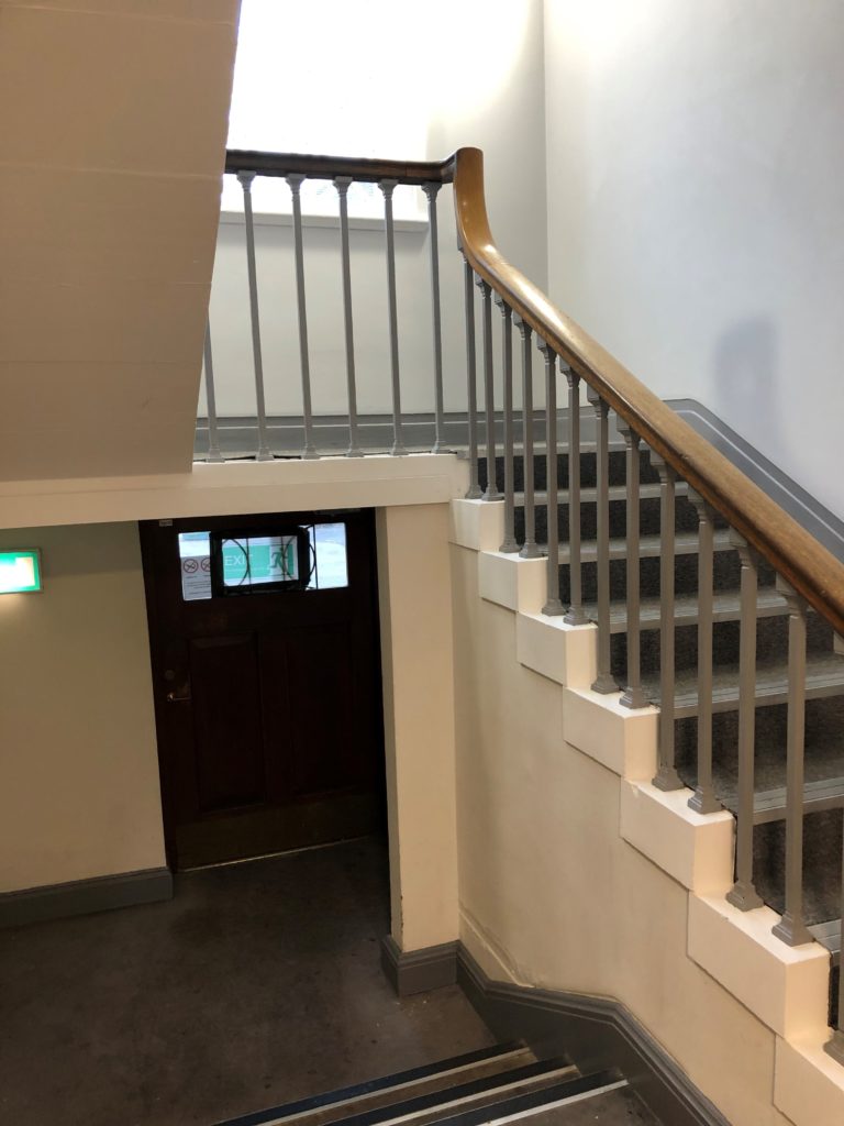 Photograph shows a stairwell with the stairs to the right and a small wooden door to the left, underneath the half-landing of the stairs.