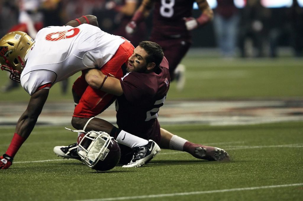 Photo from an American football game. A male player tackles an opponent while noticing that the helmet has slipped off his head.