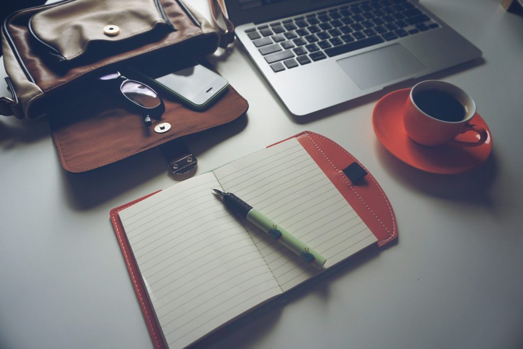 Desk space with cup of coffee, note pad, pen, laptop, handbag, glasses and phone