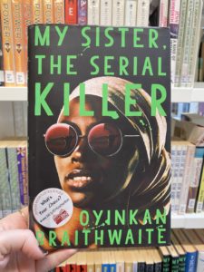 A book is held up with books on shelves visible in the background. The front cover of the book features green writing that reads, My Sister the Serial Killer, Oyinkan Braithwaite, behind which is an image of a black woman wearing a headscarf and round red tinted sunglasses.