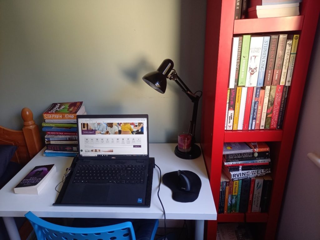 a white desk with a laptop open on it, behind is a small stack of books and a black desk lamp. to the right is a red bookshelf full of books. 