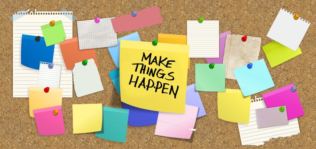 Cork board with multicoloured post it notes pinned to it. One post it note has 'make things happen' written on it.
