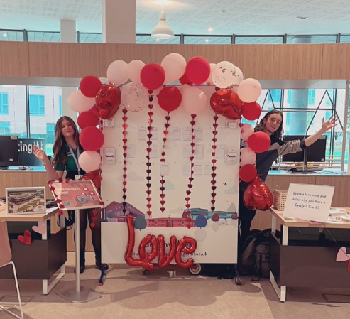 Student Advisors Rosie and Eoin staffing the Catalyst Crush stand on the ground floor of Catalyst in February 2023. They are standing either side of a feedback wall decorated with a Valentine's themed balloon arch and inflated balloon letters that spell out 'love' and there are also chains of hearts.