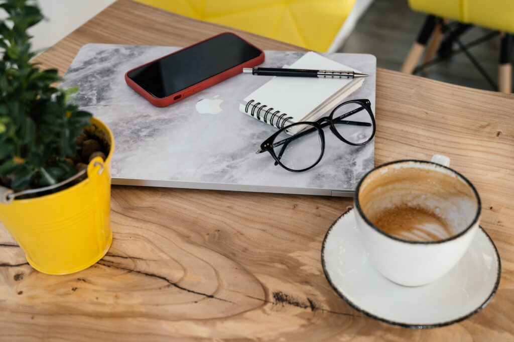 A wooden table with items on it including green plant in a yellow pot, laptop, mobile phone, pen and note pad, black rimmed glasses and an empty cup and saucer.