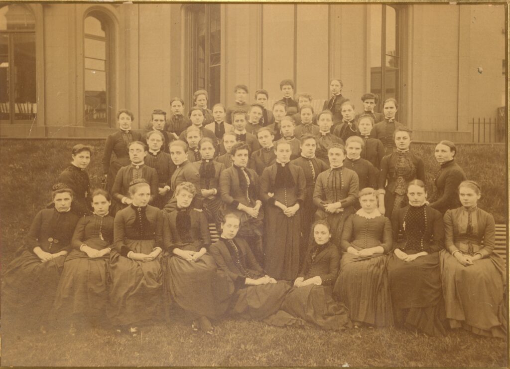 A sepia photograph of a group of women in front of a building. They are all dressed in Victorian dresses. 
