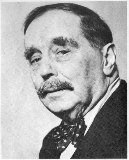 A black and white photograph of Victorian author H.G. Wells.  All that is visible are his head and upper shoulders. His head is slightly angled to the left, and he is looking towards the camera. He has dark hair and his eyebrows and moustache are bushy. He is wearing a dark jacket, a white shirt with a polka dot bow tie. 