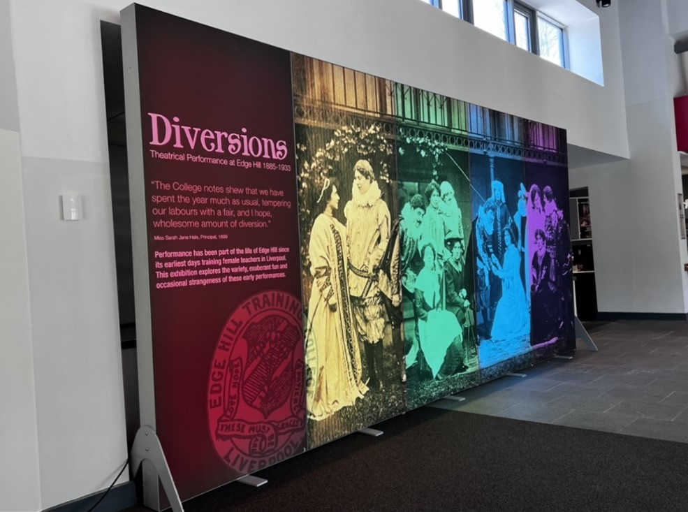 A large display board. Most of the board is a a photograph of people in Renaissance-style costumes. There is a rainbow filter placed over the top of the image. The left hand side of the board is deep pink and contains the title 'Diversions: Theatrical Performance at Edge Hill 1885-1933'. Beneath this title is the following quote from Miss Sarah Jane Hale, Principal in 1899, 'The college notes shew that we have spent the last year, as much as usual, tempering our labours with a fair, and I hope, wholesome amount of diversion.' Underneath this quote is written: 'Performance has been part of the life of Edge Hill since its earliest days training female teachers in Liverpool. This exhibition explores the variety, exuberant fun and occasional strangeness of these early performances'. At the bottom of the display is an Edge Hill University crest. 