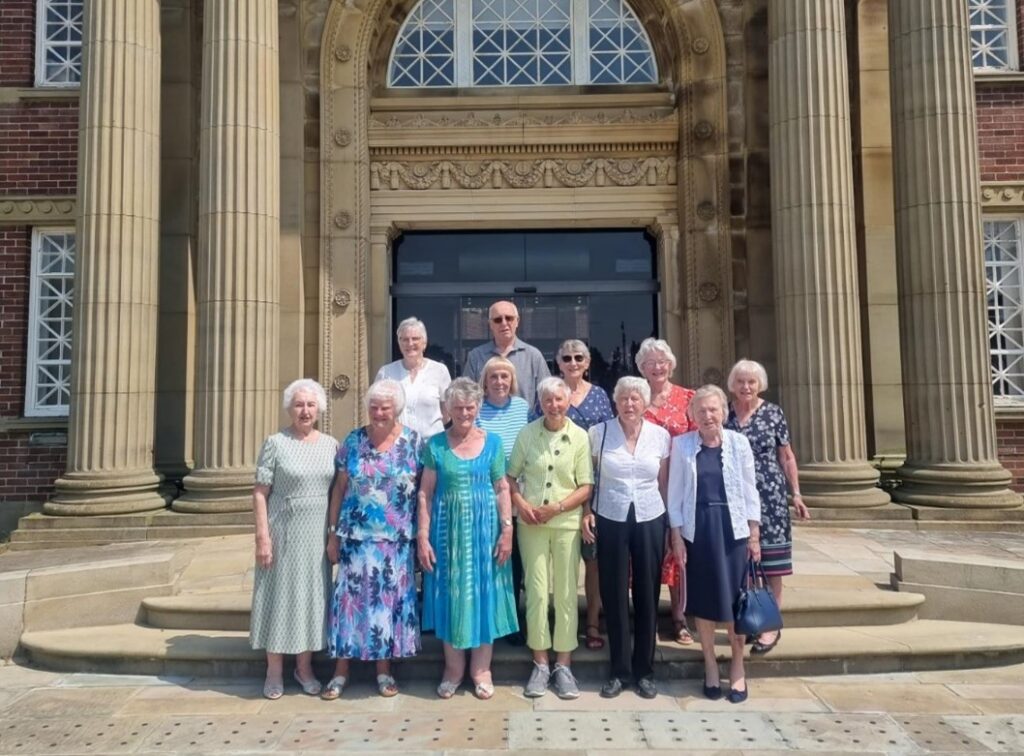 A group of twelve elderly people stood on some steps posing for a photograph. There is one male, stood at the centre back of the group, and the rest of the people are female. 