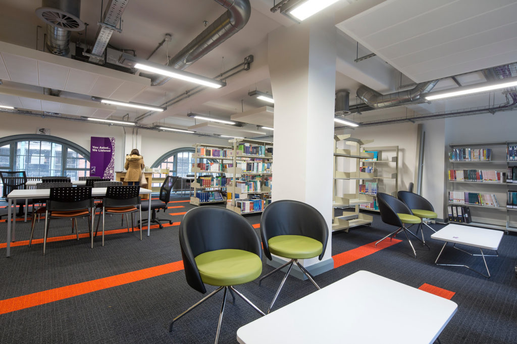 The library at St James, with seating areas and book stacks, a student checking out a book at the self-issue machine near the windows.