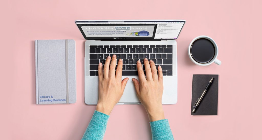 Pink table with laptop open and hands typing. To the left is a notebook, to the right is a cup of coffee, pad and pen.