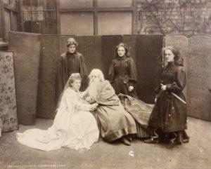 A sepia photograph featuring five female students in costume. One on the left is in a flowing white gown, crouched next to another student with a long beard. The other three students are standing and dressed in black, one in the forefront with what appears to be a fake sword.
