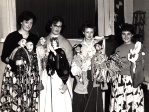 Four students in 1950s attire and standing side by side and smiling, are holding what appear to be handmade puppets on sticks.