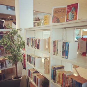 The photograph shows two white bookshelves situated from the middle of the image, coming towards the camera. On the shelves are fiction books and on the top a Black History Month display, featuring books; The Mermaid of the Black Conch and Swing Time and a framed poster. To the left of the bookshelves is a tall plant and a hint of bookshelves in an L-shape.