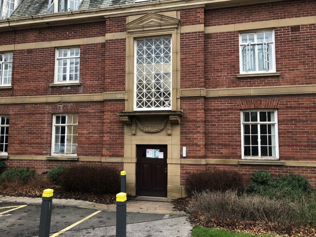 Photograph shows wall of the main building with a large vertically rectangular window which is 8 squares down and 4 across, with each square pane outlined in white with a white cross, giving a lattice appearance overall. There is a ledge and stonework underneath, and then a shortened brown wooden door which goes from the car park level to just above the hedge which runs either side in front of the doorway.