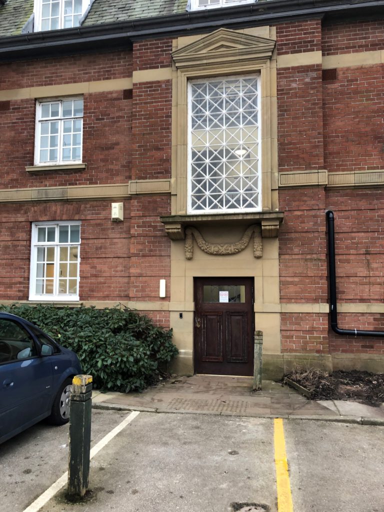 Photograph shows wall of the main building with a large vertically rectangular window which is 8 squares down and 4 across, with each square pane outlined in white with a white cross, giving a lattice appearance overall. There is a ledge and stonework underneath, and then a shortened brown wooden door which goes from the car park level to just above the hedge which runs either side in front of the doorway.