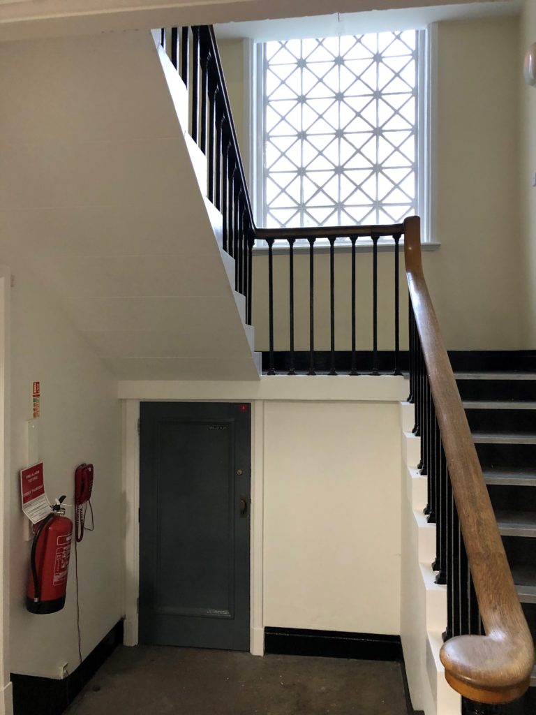 Photograph shows a stairwell with the stairs to the right and a small wooden door to the left, underneath the half-landing of the stairs.