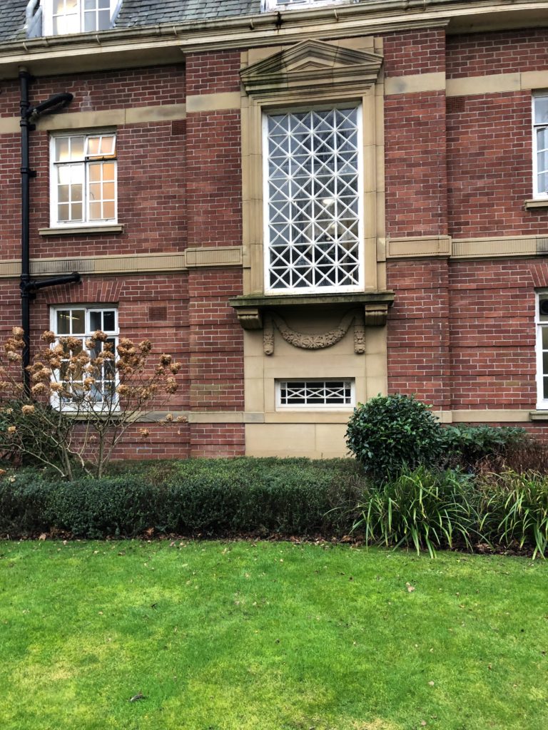 Photograph shows the green lawn in the foreground, a darker green hedge behind, and then the wall of the main building. In the centre of the wall there is a large vertically rectangular window which is 8 squares down and 4 across, with each square pane outlined in white with a white cross, giving a lattice appearance overall. There is a ledge and stonework underneath, and then closer to the ground a small horizontal window which is very thin, only 3.5 squares across and 1.5 squares down.