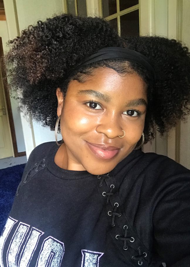 Judea, wearing a black top, hoop earrings and a head band, smiling at the camera