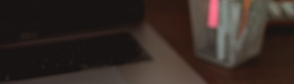 A laptop next to a pot of pens, with a notebook and various other stationery laid on top