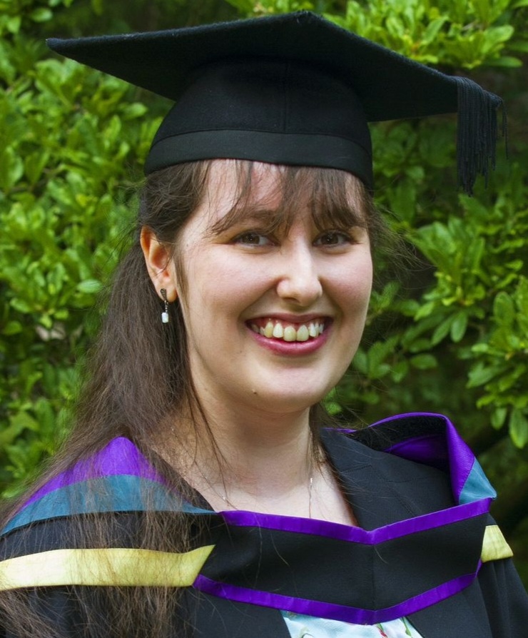 Headshot of a woman in a graduation cap and gown stood in front of a green hedge. 