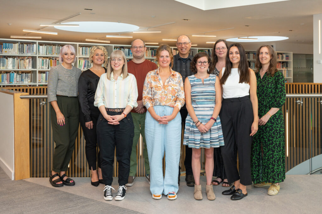 A group photo of the Student Engagement team smiling and standing together in front of the bookshelves in Catalyst.