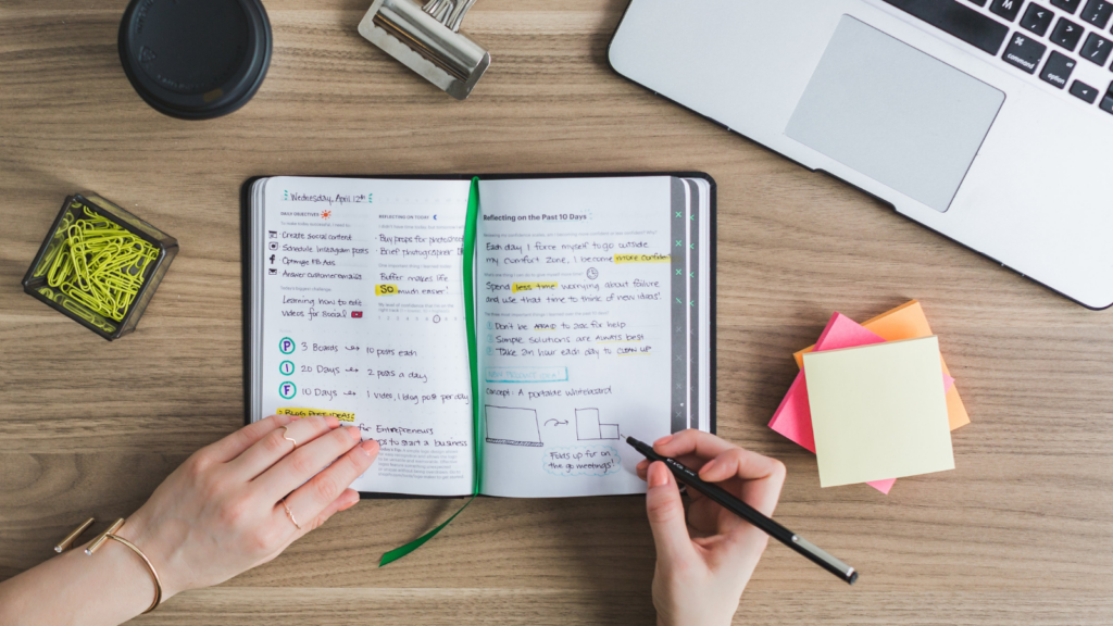 Desk with various study items on it including laptop, bulldog clip, coffee cup, sticky notes and tub with paper clips in. Someone is also writing in a notepad. You can only see their hands and they are holding a pen.