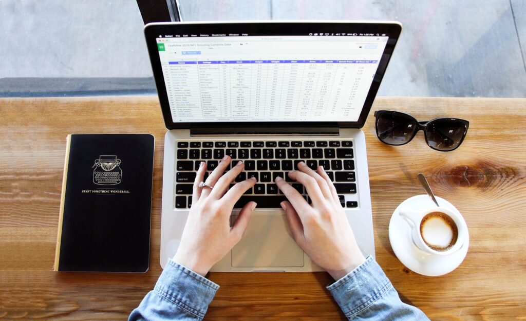 Desk with someone typing on a laptop. Also on the table is a cup of coffee, sunglasses and a notebook.