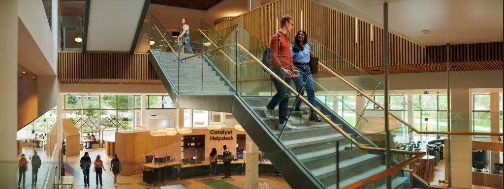 Photo of the interior of Catalyst taken from the mezzanine. Catalyst Helpdesk is in the background. In the foreground is the main staircase going from the ground floor up to the first floor and two students are walking down the stairs. 