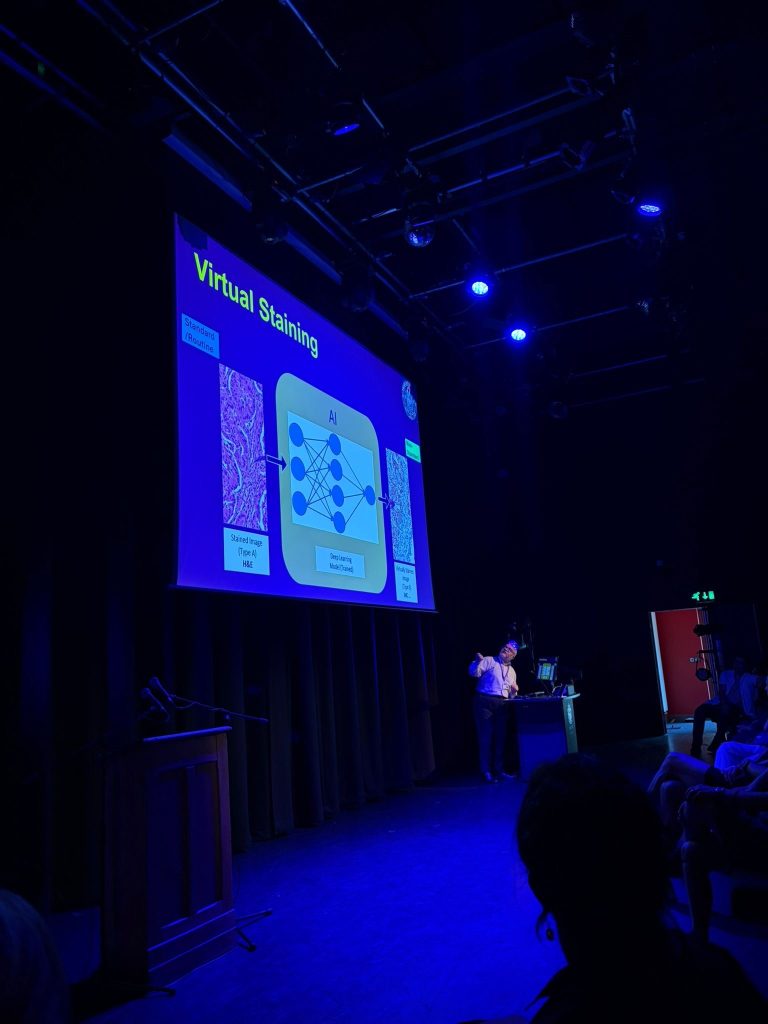 A large projector screen showing a powerpoint slide with a presenter in front of it. At the Research showcase and celebration 