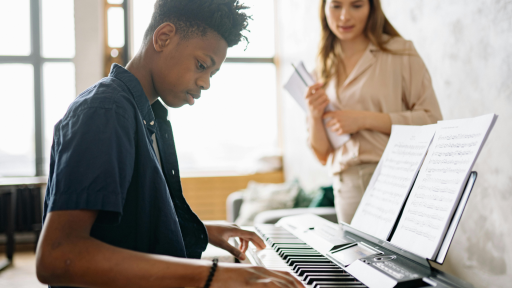 A boy is sat at a piano having a lesson. A female teacher is stood in the background watching him.