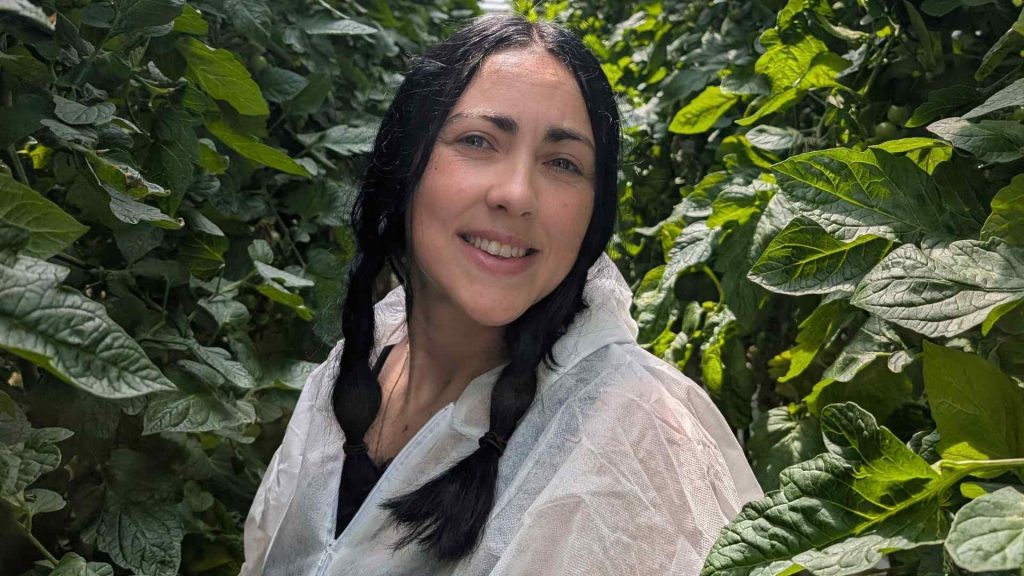 A picture of a girl looking at the camera and smiling. She is in a hazmat suit and is in a greenhouse.