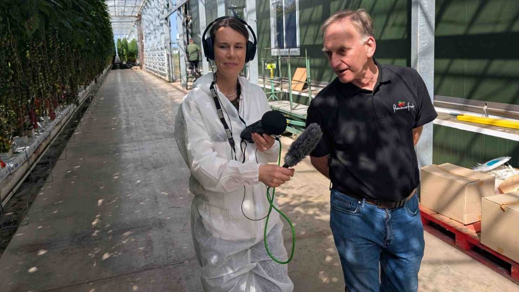 A man in a Flavourfresh t shirt is talking to a reporter who is in a hazmat suit. She has a microphone pointing at him. 